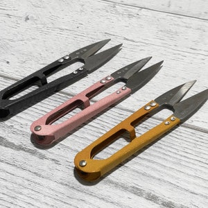 Puede incluir: Tres pares de pequeñas tijeras de tela con mangos negros, rosas y dorados. Las tijeras tienen una punta puntiaguda y están diseñadas para trabajos de detalle.