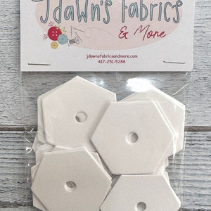 May include: A package of white hexagon-shaped fabric templates with a hole in the center. The package is labeled "Dawn's Fabrics & More" with a website and phone number.