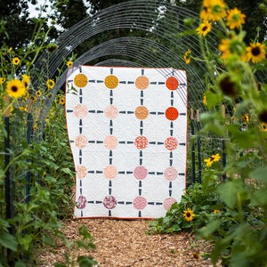 May include: A white quilt with a grid pattern of colorful circles. The quilt is displayed in a garden setting with sunflowers in the foreground.