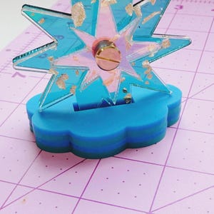 May include: A decorative star-shaped ornament made of translucent teal acrylic with embedded gold-colored flakes. The star is mounted on a layered, cloud-shaped base in shades of blue and green, secured by a central metallic screw fastener.