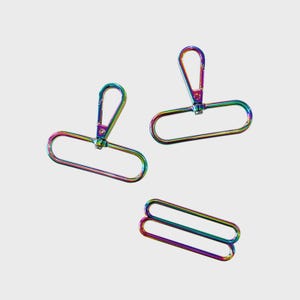 Pode incluir: Três fechos giratórios de metal com cores do arco-íris e dois deslizadores retangulares correspondentes. Os fechos têm um design de garra de lagosta com uma base de anel em D. O hardware é feito de metal brilhante e iridescente.
