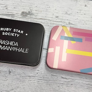 May include: Two black metal tins with white text. The tin on the left has the text "Ruby Star Society" and "Rashida Coleman Hale". The tin on the right has a pink background with a colorful abstract pattern of lines and shapes.
