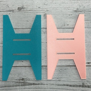 Könnte beinhalten: Zwei Pappständer, einer türkis und einer hellrosa, mit einem einfachen Design. Beide Ständer haben zwei horizontale Linien, die in die Vorderseite geschnitten sind.