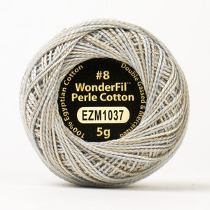 Könnte beinhalten: Ein Knäuel WonderFil Perle Cotton Garn in Größe #8 in hellgrau und beige. Das Knäuel ist mit dem Markennamen, der Garnstärke und dem Gewicht gekennzeichnet. Es besteht aus 100% ägyptischer Baumwolle, ist doppelt gekämmt und mercerisiert. Das Knäuel wiegt 5 Gramm.