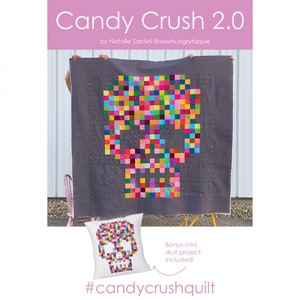 Puede incluir: Un patrón de colcha de calavera pixelada de colores sobre un fondo gris. La colcha se muestra en un soporte con una versión de almohada más pequeña del diseño de la calavera. El texto "Candy Crush 2.0" y "#candycrushquilt" están incluidos en la imagen.