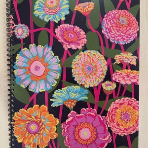 Könnte beinhalten: Spiralgebundenes Notizbuch mit einem lebendigen Blumendesign. Das Cover zeigt bunte Zinnien in Rosa, Orange, Blau und Gelb vor einem dunklen Hintergrund mit grünen Blättern und rosa Stielen. Das Notizbuch hat eine schwarze Spiralbindung.