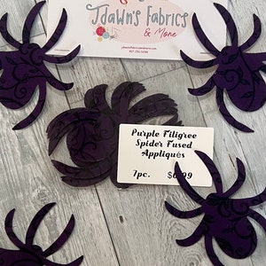 May include: Seven purple spider appliques with a black filigree design. The appliques are fused and ready to be ironed onto fabric. The text on the packaging reads "Purple Filigree Spider Fused Appliques 7pc. $6.99"