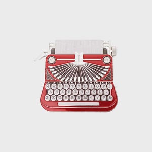 Peut inclure: Épinglette de machine à écrire rouge avec une insertion de papier blanc. L'épinglette présente un clavier détaillé avec des touches blanches et des lettres noires. Le design comprend un élément décoratif en forme d'éventail et un cadre argenté.