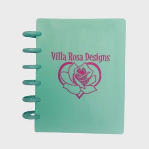 May include: A mint green disc-bound notebook featuring a dark pink logo on the front cover. The logo displays the text Villa Rosa Designs above a stylized rose graphic enclosed within a heart shape. The notebook has six circular plastic discs.