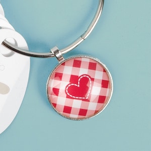 Pode incluir: Um chaveiro prateado com um charme de coração xadrez vermelho e branco. O coração tem um contorno branco costurado.