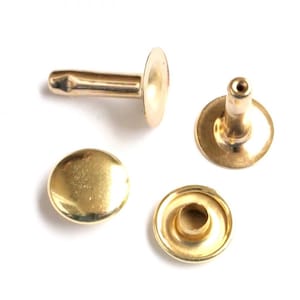 Puede incluir: Cuatro remaches de metal de color dorado, dos con la parte superior plana y dos con el centro hueco. Los remaches se utilizan para sujetar tela o cuero.