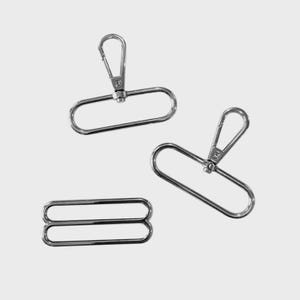 Pode incluir: Três peças de ferragens metálicas prateadas. Duas peças têm um laço retangular com um fecho giratório, e uma tem um laço retangular duplo. As ferragens são projetadas para bolsas ou correias.