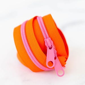 May include: A coiled zipper featuring bright orange fabric tape and contrasting pink plastic teeth. The zipper includes two matching pink pull tabs with circular openings, designed for sewing projects requiring a vibrant and functional closure.