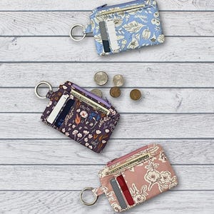 May include: Three fabric mini wallets with zippered tops and exterior card slots, each featuring a metal key ring. The wallets display floral patterns in light blue, dark purple, and dusty pink. Text includes "Fabric Mini Wallet" and "ATB-244".