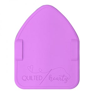 May include: A lilac-coloured, house-shaped sewing mat with two side clips. The mat features the text "QUILTED heartz" with a dotted line and heart design. The mat is designed for quilting and sewing projects.