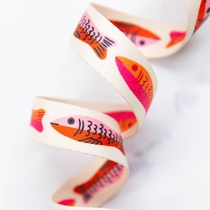 Puede incluir: Una cinta tejida de color crema con un patrón repetitivo de peces estilizados en tonos vibrantes de rosa y naranja. Los peces están representados con ojos negros simples y líneas detalladas en sus cuerpos, dispuestos en un diseño en espiral.
