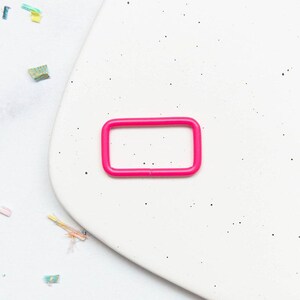 Peut inclure: Une boucle rectangulaire en métal rose vif avec des coins arrondis. La pièce de quincaillerie présente une finition lisse de couleur unie et un design de cadre continu, adaptée pour servir de connecteur de sangle ou de composant fonctionnel.