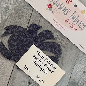 May include: Five black and white skull and crossbones fabric appliques. The appliques are fused and ready to be sewn onto a project. The appliques are shaped like a spider with a skull and crossbones pattern. The text on the card reads "Skull Filigree Spider Fused Appliques 5pc. $5.75".