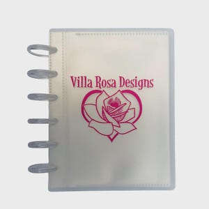 May include: A translucent disc-bound notebook featuring a frosted cover with the text Villa Rosa Designs printed in magenta above a stylized magenta rose graphic. The notebook has six clear plastic binding discs along the left side.