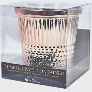 May include: A rose gold colored thimble shaped container with vertical ridges and a dimpled base. The container is enclosed in a clear plastic box with a black label that reads THIMBLE CRAFT CONTAINER and the brand name Hemline at the bottom.