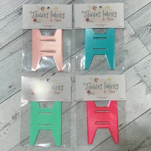 Puede incluir: Cuatro formas de artesanía de plástico en diferentes colores: rosa, turquesa, verde y rojo. Cada forma es un diseño simple de una escalera con tres peldaños. Las formas están empaquetadas en bolsas de plástico transparentes con una tarjeta de encabezado blanca que dice "Jdawn's Fabrics & More".