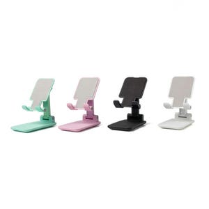 May include: Four adjustable phone stands in pastel colors: mint green, pink, black, and white. Each stand has a textured surface for device placement and a foldable design for portability. The stands are designed to hold a phone or tablet.