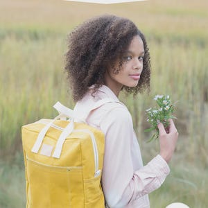 Könnte beinhalten: Ein gelber Rucksack mit weißen Trägern und einer Vordertasche, mit dem Text MAKING BACKPACK create your own backpack: regular & mini sizes, sowie einem runden Abzeichen mit der Aufschrift 2 sizes to sew! und dem Markennamen noodlehead.