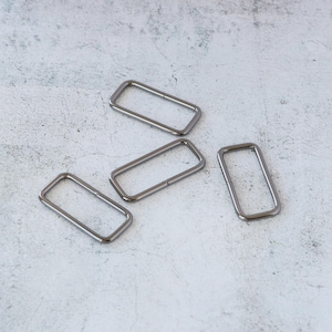 May include: Four silver-colored rectangular metal rings. The rings are evenly spaced and lie on a light gray, textured surface. The rings have rounded corners and a smooth, shiny finish. These rings are likely used for bag making or other crafting projects.