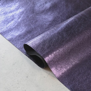 May include: A close-up of a roll of purple leather with a metallic sheen. The leather has a textured surface and a dark backing. The color transitions from a lighter purple to a deeper shade, creating a gradient effect.