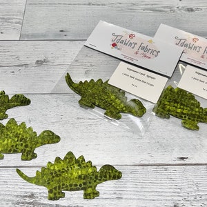 Könnte beinhalten: Vier grüne Stoff-Dinosaurier-Applikationen in einer Packung mit einem Etikett, auf dem "Dawn's Fabrics & More" und "Dinosaur Land Appliques 1 Stück pro Stück, Bügeltransfer 5,00 €" steht.