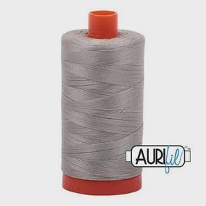 Pode incluir: Um carretel de linha de costura cinza claro enrolado em torno de um núcleo cilíndrico com tampas de plástico laranja brilhante na parte superior e inferior. A marca AuriFil está impressa em preto numa etiqueta branca na lateral.