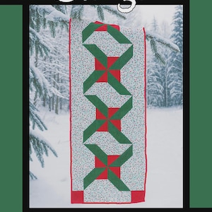 May include: A Christmas-themed quilt with a green and red geometric design, hanging from a snowy tree branch. The word "Jingle" is at the top. Designed by Tricia Lynn Maloney for Villa Rosa Designs.
