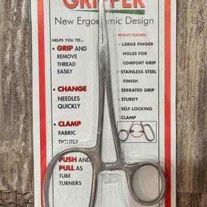May include: A stainless steel Super Gripper hemostat tool with a self-locking clamp and serrated grip, packaged on a white card with red borders. The packaging features text including The Super Gripper, New Ergonomic Design, and product features.