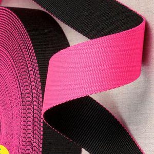 Puede incluir: Un rollo de cincha de nailon negra y rosa intenso. La cincha está tejida apretadamente y parece duradera. La sección rosa intenso está doblada sobre la sección negra, mostrando el contraste de color. El rollo está en el lado izquierdo de la imagen.