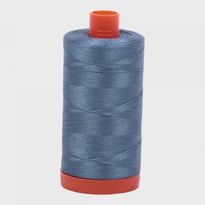 Puede incluir: Un carrete de hilo de coser color azul pizarra enrollado firmemente alrededor de un núcleo cilíndrico. El carrete presenta una tapa de plástico naranja brillante en la parte superior y una base de plástico naranja a juego en la parte inferior.