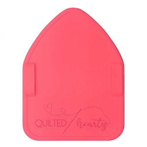 May include: A coral-colored, house-shaped pressing mat with two attached side straps. The mat features the text "QUILTED heartz" with a heart and dotted line design. The mat is designed for quilting and sewing projects.