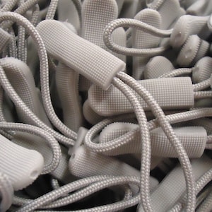 May include: A pile of gray drawstring cord stoppers. The stoppers are made of a soft, textured material and have a simple, functional design.