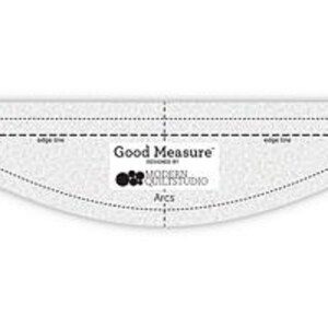 May include: A clear, arc-shaped quilting ruler with dotted and solid lines for measuring and cutting fabric. The ruler has the text "Good Measure" and "Designed by Modern Quilt Studio" printed on it. The tool is used for quilting.