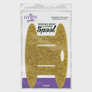 May include: A gold glittery spool for binding wrap and store, packaged in a clear plastic bag. The packaging features the text "the GYPSY quilter", "ORGANIZE YOUR QUILT BINDINGS IN STYLE", "BINDING WRAP & STORE Spool", and "1 COUNT" in purple and black.