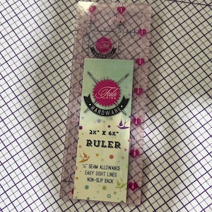 Puede incluir: Una regla de plástico transparente con un diseño de corazón rosa. La regla mide 5,1 cm por 15,2 cm y tiene una tolerancia de costura de 0,6 cm. También tiene líneas de visión fáciles y una parte posterior antideslizante. La regla está etiquetada como "Tula Pink Hardware".