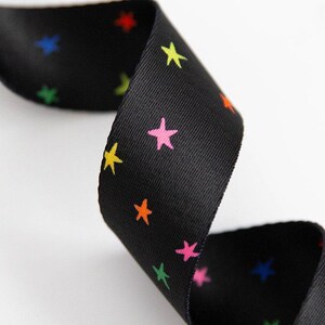 Puede incluir: Cinta negra con un patrón repetitivo de estrellas coloridas en amarillo, rosa, naranja, verde, azul y rojo. La cinta está rizada, mostrando el patrón en ambos lados. El fondo es blanco.
