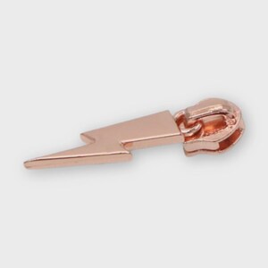 May include: A rose gold zipper pull in the shape of a lightning bolt. The pull is made of metal and has a smooth, shiny finish. The lightning bolt design is a popular symbol.