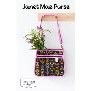 Könnte beinhalten: Eine schwarze, gesteppte Handtasche mit floralem Muster und rosa Einfassung, die an einem Holzgestell hängt. Die Tasche hat einen langen rosa Riemen und ist mit Blumen gefüllt. Der Text "Janet Mae Purse" steht oben.
