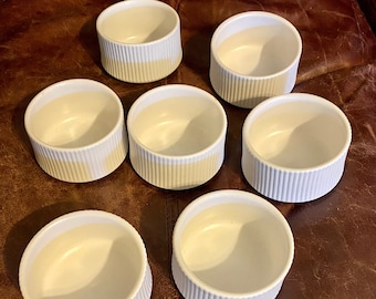 May include: Seven white ceramic ramekins with a light yellow band around the top. The ramekins are arranged in a circle on a brown leather surface.