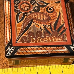May include: A square ceramic box featuring a hand-painted bird and floral motif in shades of orange, black, and brown. The lid displays a central bird illustration with geometric borders. A yellow measuring tape shows the box is 4 inches wide.