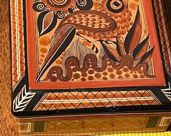 May include: A square ceramic box featuring a hand-painted bird and floral motif in shades of orange, black, and brown. The lid displays a central bird illustration with geometric borders. A yellow measuring tape shows the box is 4 inches wide.