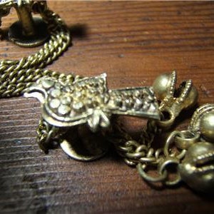 May include: Close-up of antique gold-colored jewelry. The jewelry includes a chain, decorative clasps, and small bell-shaped charms. The pieces are arranged on a wooden surface, showcasing the intricate details and aged appearance of the metal.