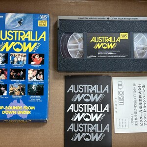 May include: A VHS video cassette tape with a black and silver case. The front of the case features a colorful image of a band and the text "Australia Music Now Video". The back of the case features a list of songs and the text "Up-Sounds From Down Under".