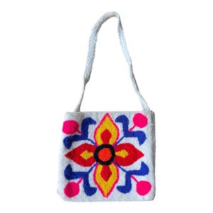 May include: White square handbag with a braided strap. The bag features a colourful floral design in red, yellow, blue, and pink. The flower has a black centre and is centered on the bag.