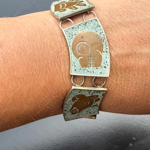 May include: Silver-toned bracelet with three rectangular panels. Each panel features a copper-colored design on a speckled background. The designs include a skull, a bird, and a deer. The bracelet is connected by silver rings.